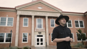 Poet Laureate Bobby LeFebre in black hat and clothes stands and speaks in front of Adams State's Richardson