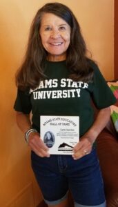 A person wearing an Adams State University t-shirt smiles and holds a plaque that says: "Adams State Educators Hall of Fame. Carey Sanchez"
