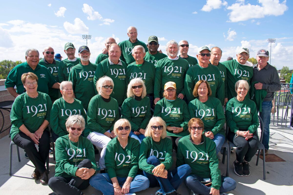 A group opf alumni stand and smile wearing green Adams State "1921" t-shirts
