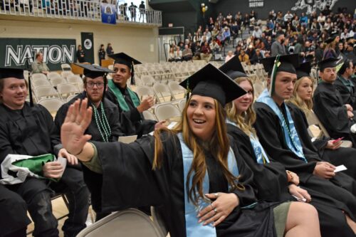 An Adams Stater waves and smiles while waiting to graduate with other students.