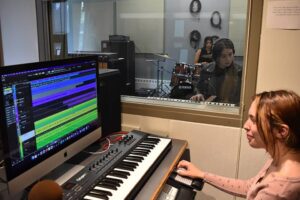 Three young musicians recording in a studio. One is mixing sound on a computer and keyboard controller, and the other is playing drums and keyboard in the sound booth.
