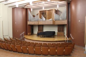 A medium-sized concert hall or music recital auditorium. The stage is a wooden semi-circle with a black grand piano centered on it. Behind the piano is a large, elaborate pipe organ with both exposed silver pipes and wooden casing. The surrounding walls are a warm reddish-brown color, and the audience seating consists of rows of curved, light-brown wooden chairs.