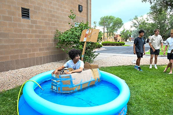 Adams State University mechanical engineering new student orientation student in cardboard homemade boat in kiddie pool testing floatation